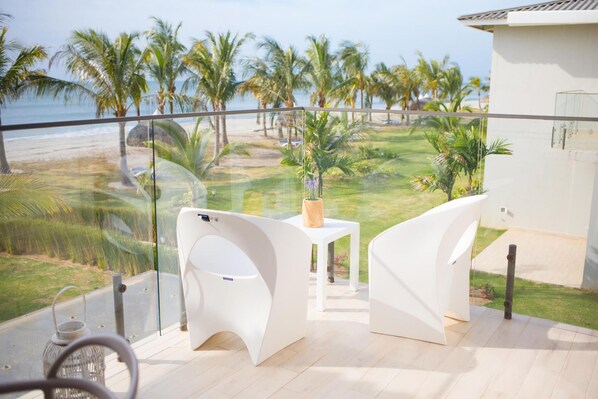 A balcony with a view of the beach and palm trees, featuring white chairs and a small table with a plant.