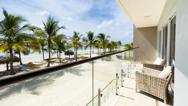 Un balcon doté d’une balustrade en verre, donnant sur une plage bordée de palmiers et surplombant un ciel dégagé.