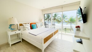 Une chambre moderne avec un grand lit, une table de chevet avec une lampe, un téléviseur fixé au mur et une vue sur la plage grâce à de grandes fenêtres.