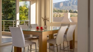 A dining area with a glass table, white chairs, and a view of mountains through large windows.
