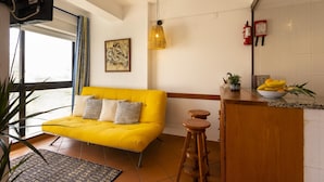 A bright yellow sofa with patterned cushions, a wooden stool, a small potted plant, and a fruit bowl on a kitchen counter. - A Torre by Acasadascasas (Mafra)