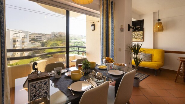 A dining area with a table set for two, a yellow sofa, and a view of the cityscape. - A Torre by Acasadascasas (Mafra)