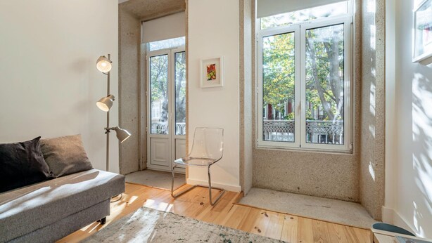 Una sala de estar moderna con un sofá gris, una silla transparente, una lámpara de pie y ventanas amplias con vistas a árboles.