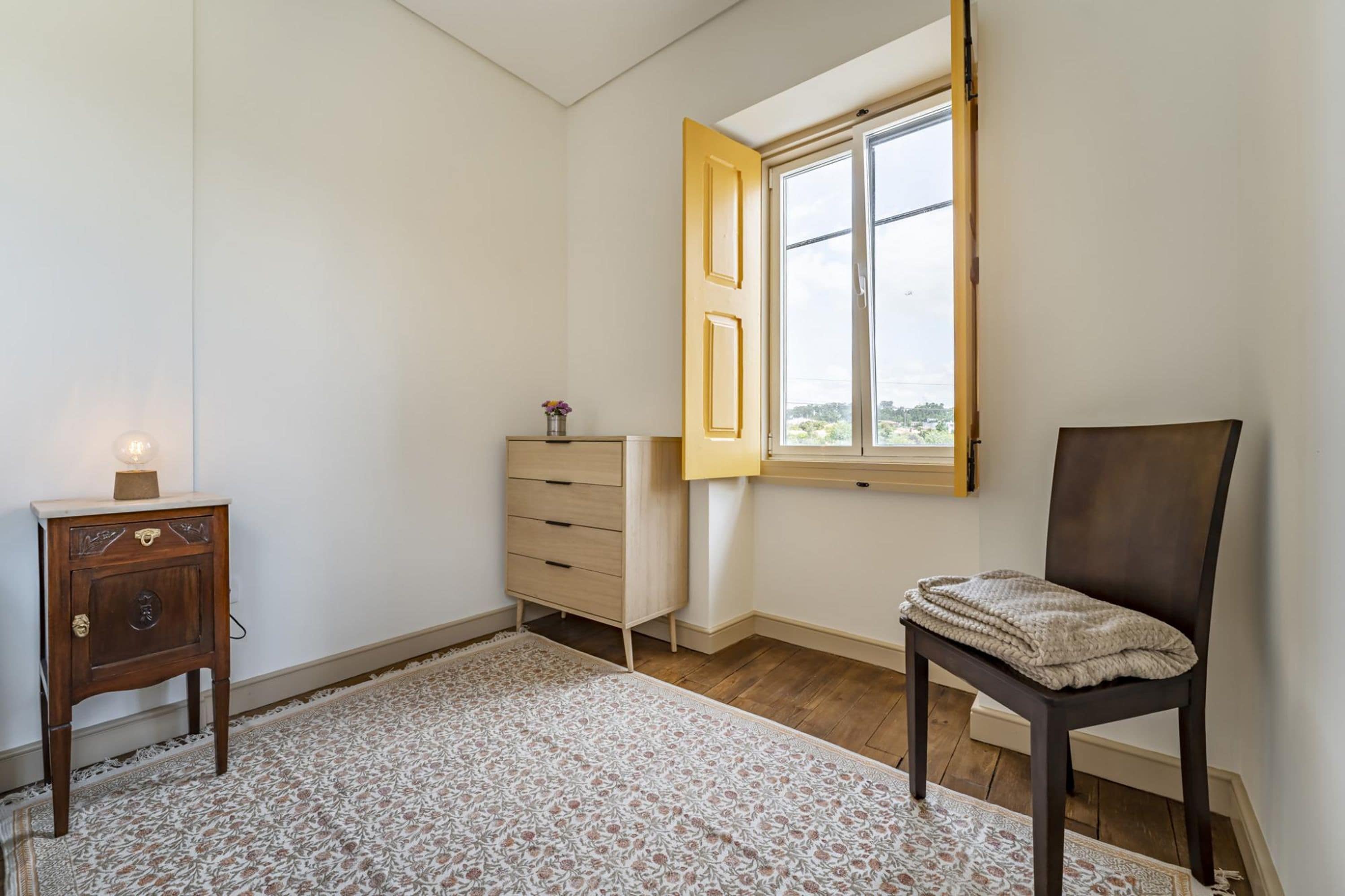 A room with a wooden floor, a patterned rug, a wooden cabinet, a chair, and a window with an open door.