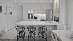 A modern kitchen with white cabinetry, a central island with bar stools, and recessed ceiling lighting. - Belva Blue in Glifada (Glyfada)