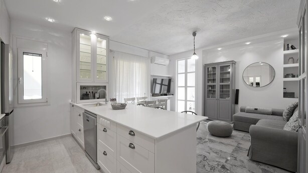 A modern kitchen with white cabinetry, a central island, and a dining area with a grey sofa and ottoman. - Belva Blue in Glifada (Glyfada)