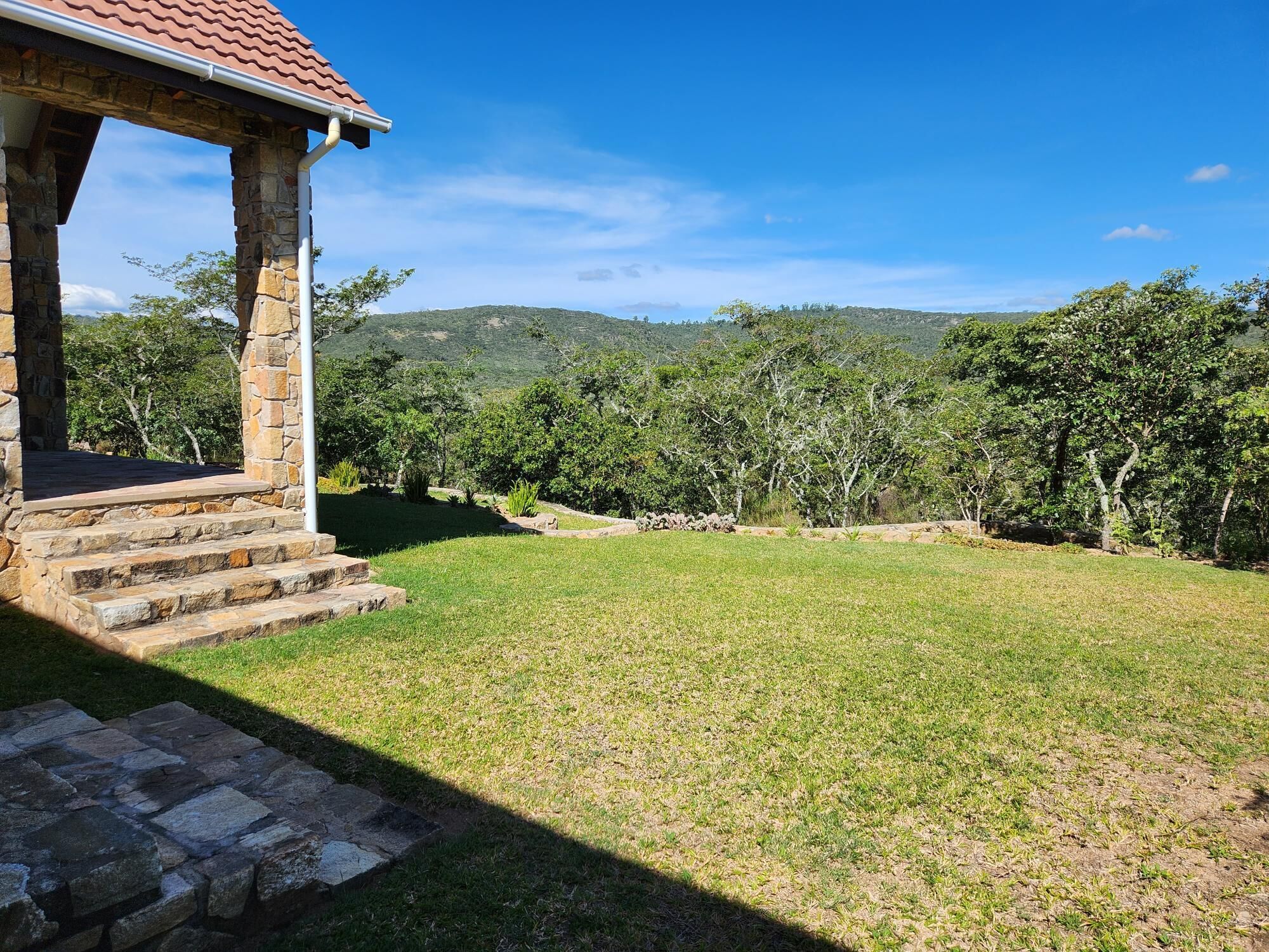 A stone gazebo with a tiled roof overlooking a grassy area with trees and hills in the background.