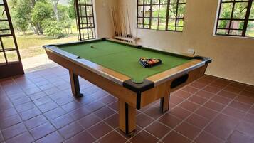 A pool table with a green felt surface, surrounded by brown tiled flooring, and windows with a view of greenery outside.