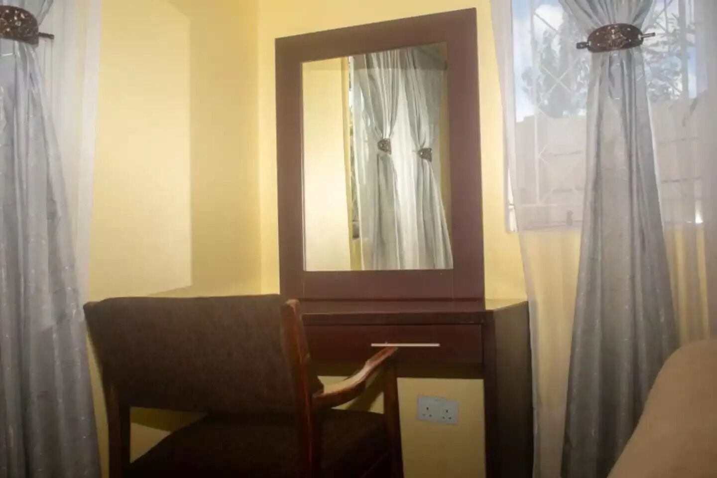 A room with a wooden desk, a chair, a mirror, and a window with sheer curtains.