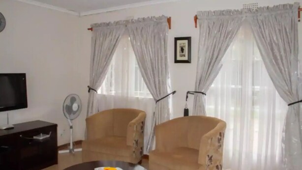 A living room with two armchairs, a TV, a fan, and a table with fruit. - Cosy 3 Bedroom Cottage - 2034 (Harare)