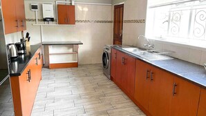 A kitchen with wooden cabinets, a washing machine, a sink, and a window with decorative elements. - Chic Modern House on Neasden - 2085 (Bulawayo)