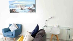A modern living room with a blue armchair, a glass coffee table, a white desk with a lamp, and a large painting of a coastal scene. - Chic Modern House on Neasden - 2085 (Bulawayo)