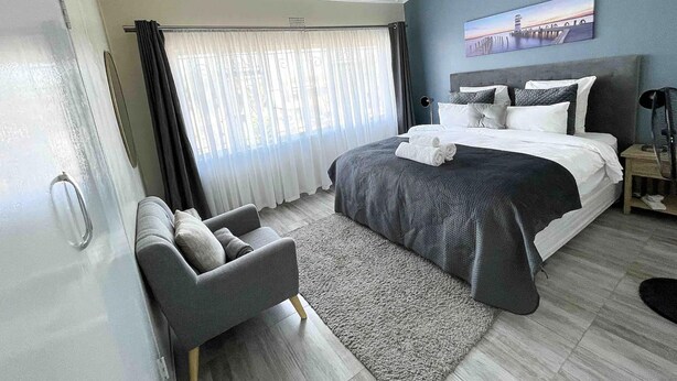 A modern hotel room with a large bed, a grey armchair, a desk, and a painting on the wall. - Chic Modern House on Neasden - 2085 (Bulawayo)