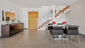 A modern hotel lobby with a reception desk, a staircase, and a dining area with chairs and a table.