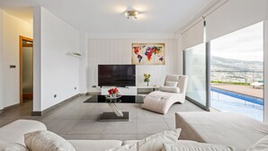 A modern living room with a large flat-screen TV, a comfortable sofa, a coffee table with flowers, and a view of a swimming pool through large windows.