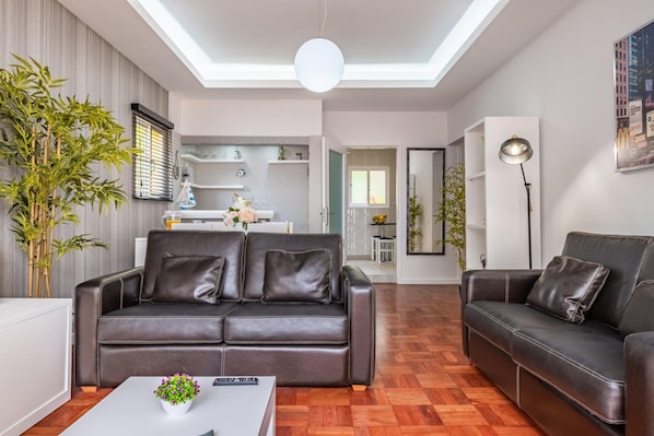 A modern living room with a brown leather sofa, a white coffee table with a plant, and a dining area in the background.