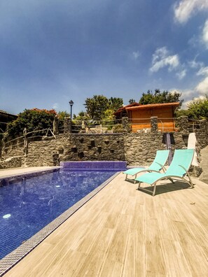 A swimming pool with a wooden deck, stone walls, and a wooden building in the background.