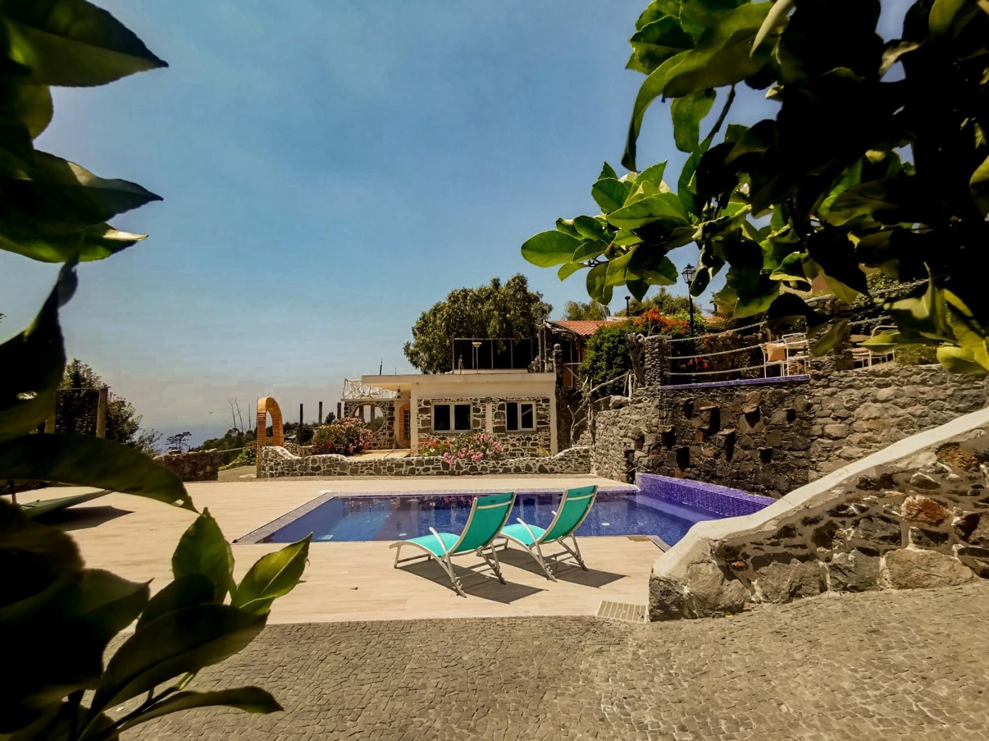 Un área de piscina con dos sillas reclinables, un muro de piedra y un edificio al fondo.