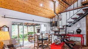 A spacious living area with a wooden ceiling, a fireplace, a dining table with chairs, a red coffee table, and a staircase. - Ivy Cottage by Homie (Santana)