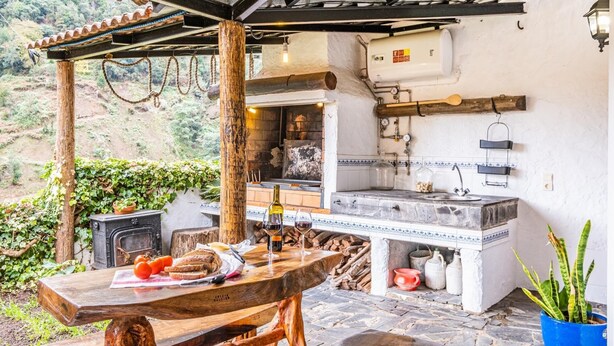 An outdoor kitchen with a stone countertop, a built-in oven, and a rustic wooden table with food and drinks. - Ivy Cottage by Homie (Santana)