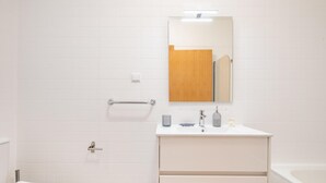 Une salle de bain avec un mur recouvert de carreaux blancs, une porte en bois, un lavabo blanc avec un miroir au-dessus, et une baignoire.
