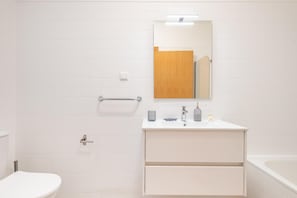 Kylpyhuone, jossa on valkoisilla laatoilla verhoiltu seinä, puuovi, valkoinen pesuallas, jonka yläpuolella on peili, ja kylpyamme.