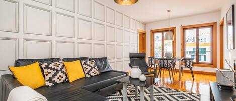 A modern living room with a black leather sofa, patterned cushions, a glass coffee table, and a geometric rug.