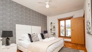 Une chambre à coucher avec un papier peint à motifs zébrés, un lit blanc avec de nombreux coussins, une table de chevet avec une lampe, un plancher en bois, une grande fenêtre donnant sur des immeubles et un ventilateur de plafond.