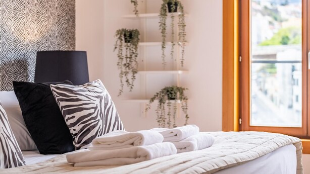 Un lit bien fait, avec du linge de lit blanc, un coussin à motifs zébrés et un coussin noir. Des plantes suspendues sont disposées sur des étagères le long du mur, et une fenêtre offre une vue sur les bâtiments.