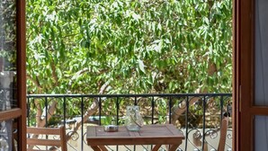 A balcony with a wooden table and chairs, a metal railing, and a view of green foliage. - Apartments for 3 1st Floor (Malevizi)