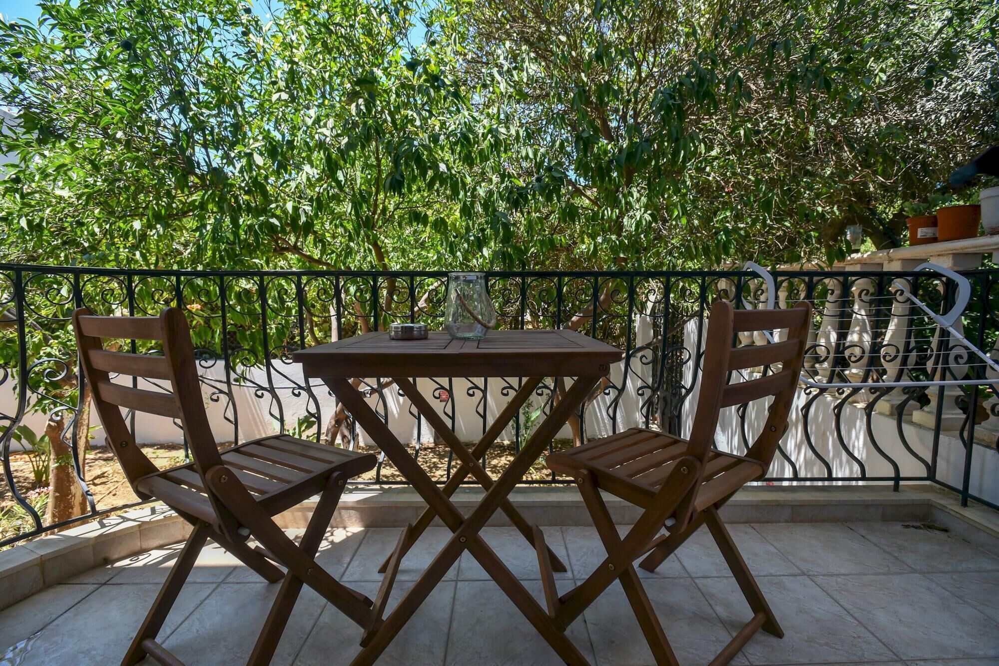 A small outdoor patio with a wooden table and two chairs, surrounded by a metal railing and lush greenery.