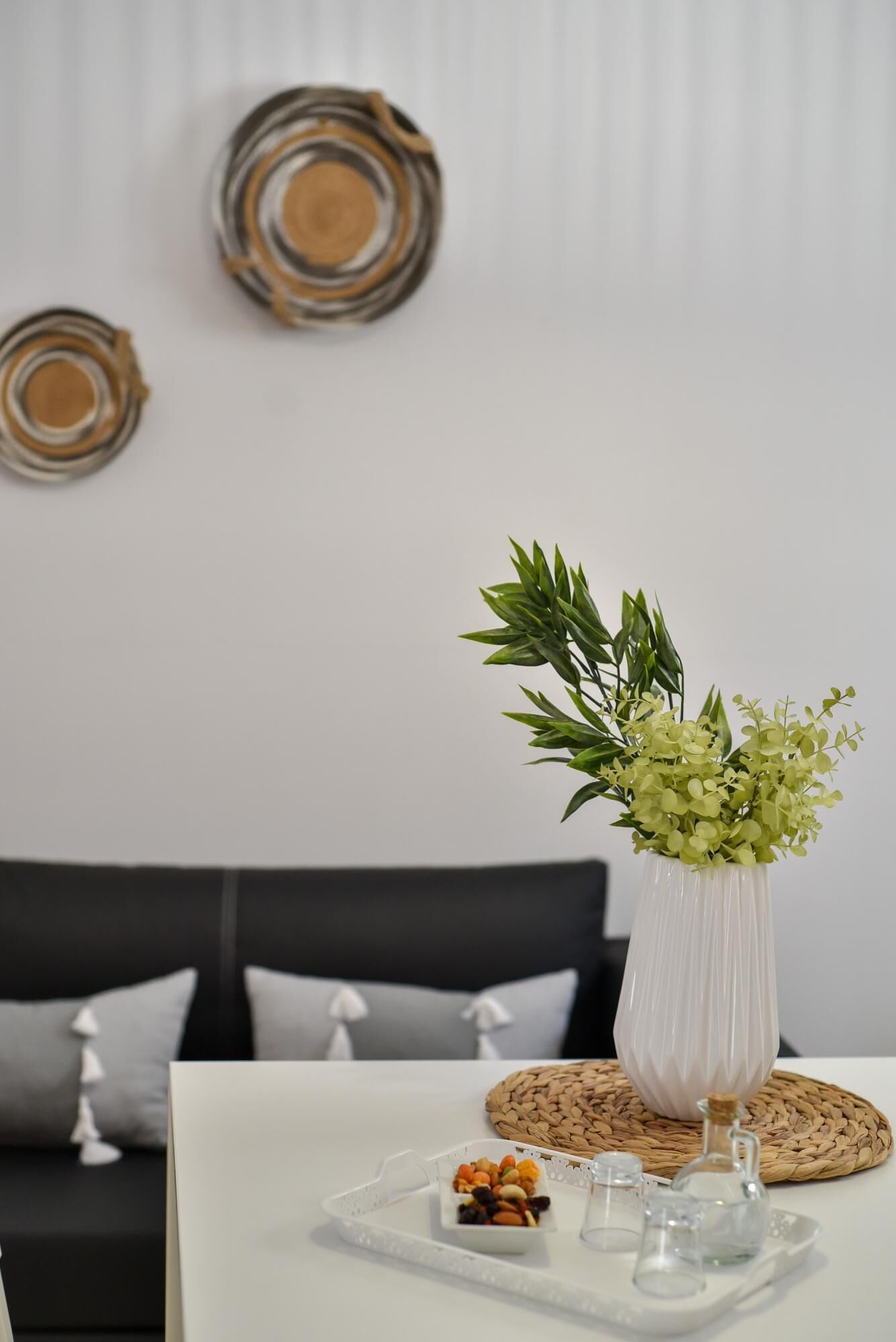 Une table à manger ornée d’un vase blanc contenant des feuilles vertes, d’un set de table en fibres naturelles et d’un plateau avec des en-cas et des verres.