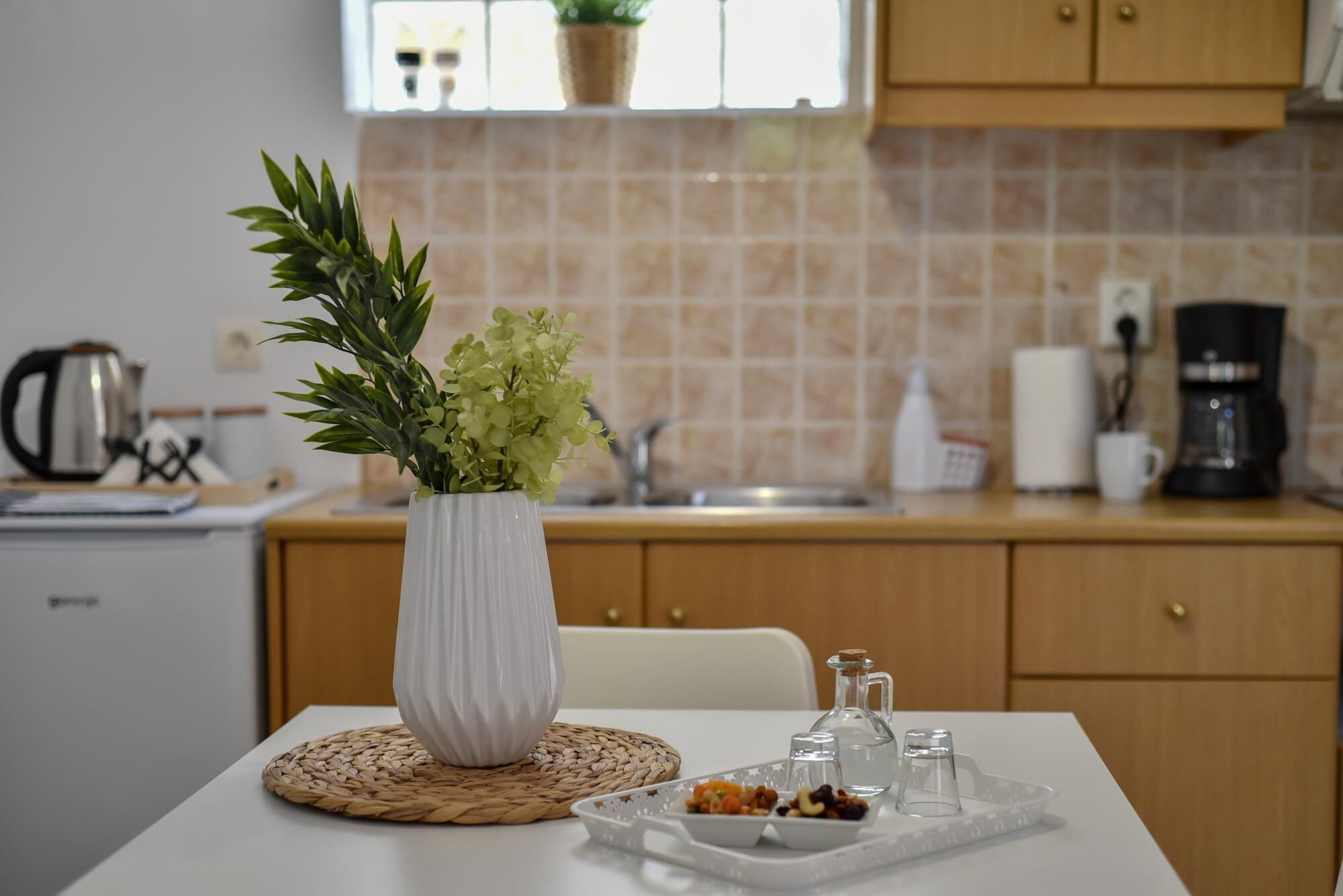 Uma cozinha com uma mesa branca, um jogo americano tecido, um vaso branco com plantas verdes e uma bandeja com nozes e frutas secas.