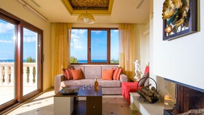 A spacious living room with a large window offering a view of the sea, a sofa with cushions, a coffee table with a decorative bowl, and a statue on the mantel. - Sunshine Villa (Hersonissos)