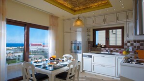 A modern kitchen with white cabinets, a stainless steel oven, and a dining area with a glass table and chairs. - Sunshine Villa (Hersonissos)