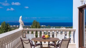 A balcony with a table and chairs, a statue, and a view of the sea and distant buildings. - Sunshine Villa (Hersonissos)