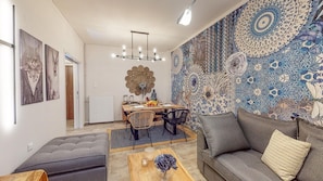 A modern living room with a dining area, featuring a patterned wall, a sofa, a coffee table, and a dining table with chairs. - Motley Elegant Apartment by Estia (Heraklion)