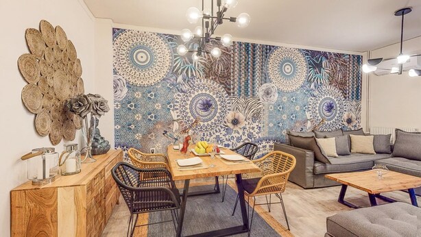 A modern living room with a dining area, featuring a large decorative tapestry, a chandelier, and a grey sofa.