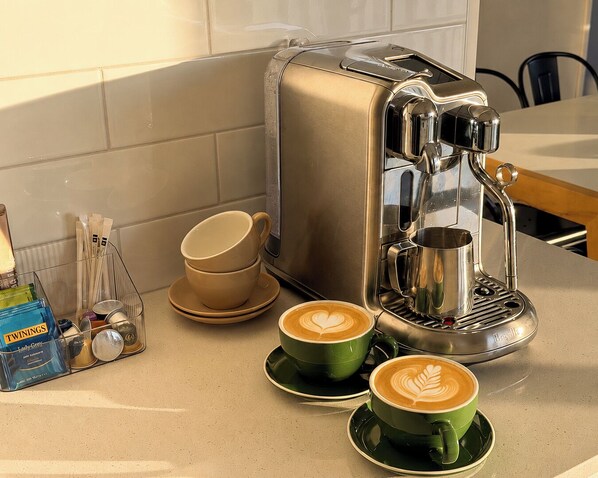 Una cocina con una máquina de café, dos tazas de café, una pila de tazas y una caja de café Twinings.