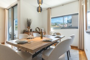 A dining area with a long table set for six, a bottle of wine, and a view of a town through large windows. - Funchal Frui Vita by Atlantic Holiday (Funchal)