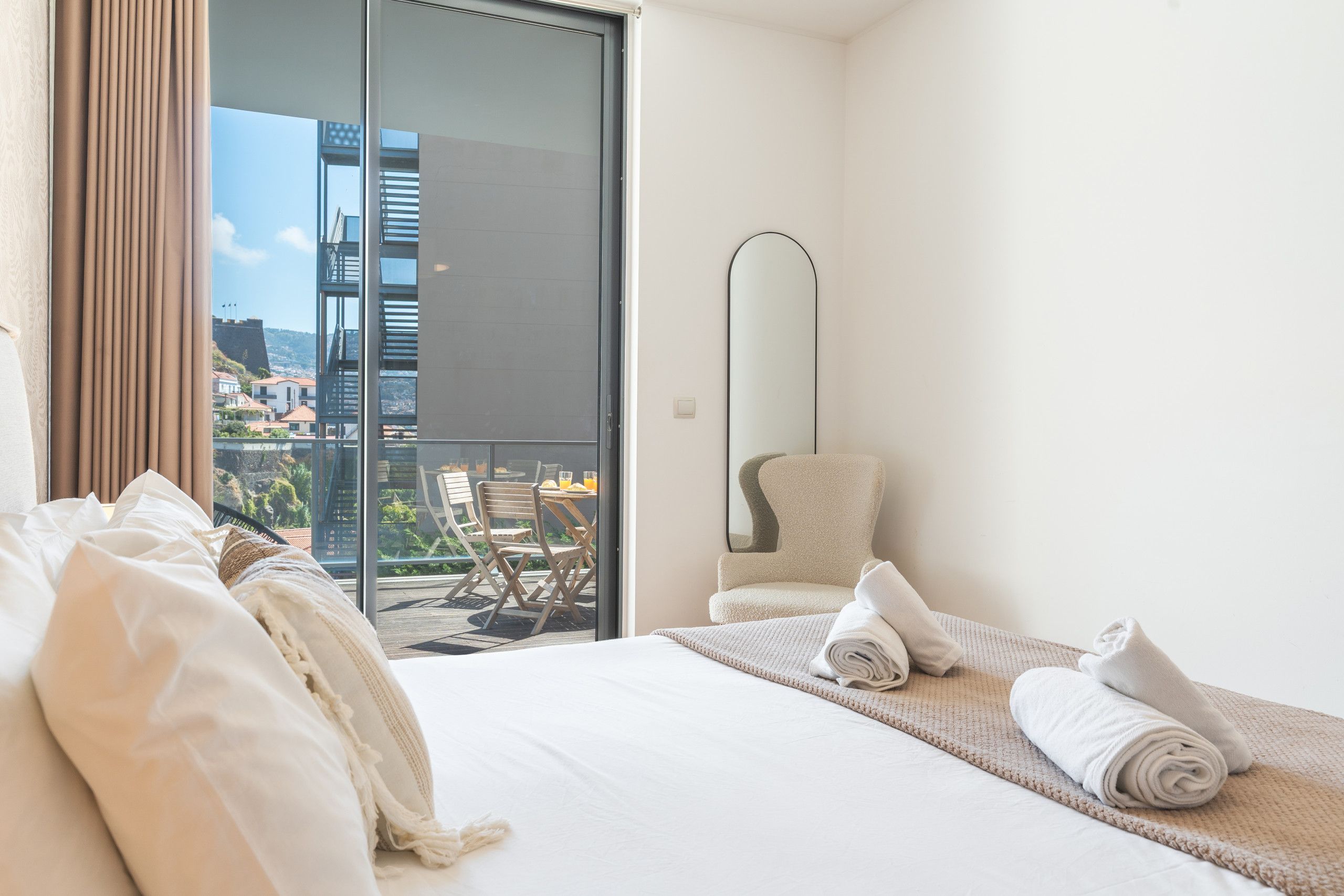 A hotel room with a bed, a chair, a large mirror, and a view of the city through a glass door.