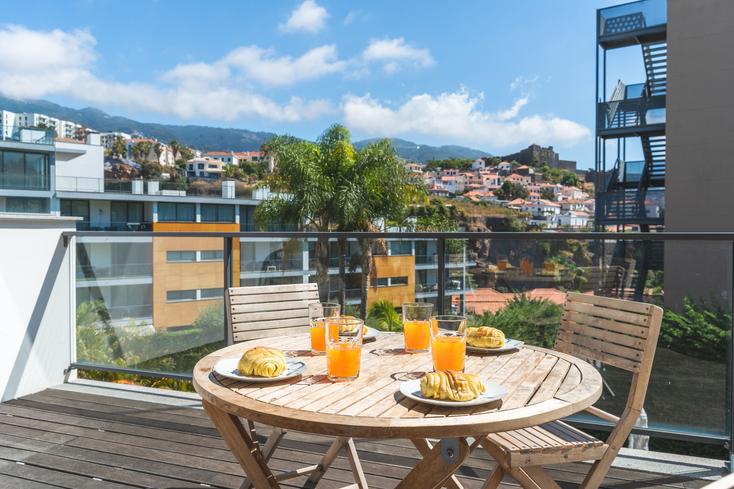 Een houten tafel met stoelen op een balkon met uitzicht op een stad, gebouwen en een berg in de verte.