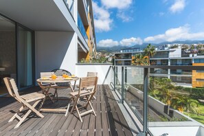 A balcony with a wooden table and chairs, overlooking a cityscape with buildings and mountains.