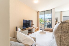 A modern hotel room with a large bed, a flat-screen TV, a small table, and a view of a cityscape through large windows.