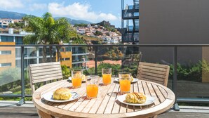 Une table en bois, sur laquelle sont disposées deux assiettes de croissants et des verres de jus d’orange, est placée sur un balcon donnant sur une ville pittoresque située sur une colline.