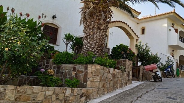 A street with a white building featuring stone walls, a palm tree, and a parked motorcycle. - Nikos Studio 2 in Gialiskari (Ikaria)