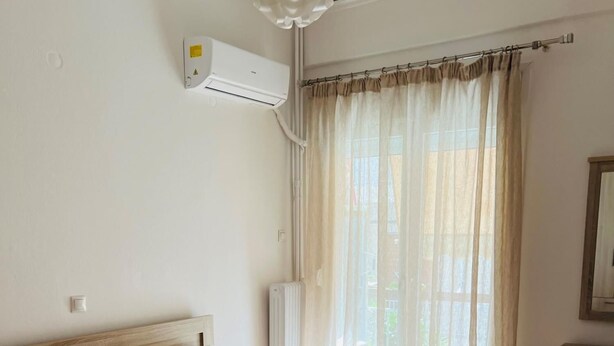 A neatly made bed with white linens and two rolled towels, a wooden headboard, a bedside table, a small mirror, and a window with sheer curtains. - Piraeus 3 in Piraeus (Piraeus)