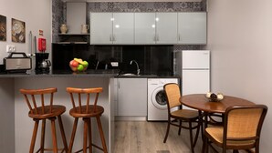 A compact kitchen with a washing machine, a coffee maker, and a dining area with two wooden chairs and a table. - Vila do Largo B by Homie (São Vicente)
