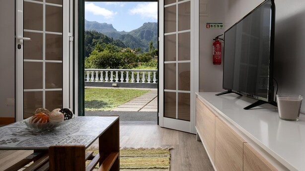 A room with a TV, a table with a bowl of pastries, and a view of mountains through an open door. - Vila do Largo B by Homie (São Vicente)