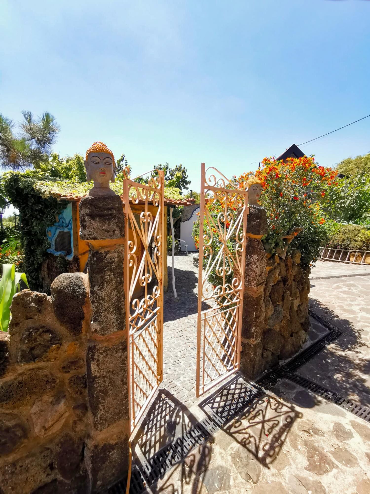 A stone gate with intricate metalwork, leading to a building with a tiled roof and surrounded by greenery and flowering plants.